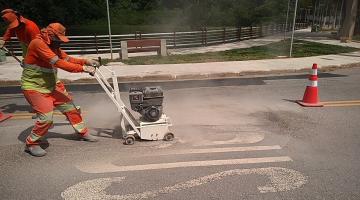 Homem utiliza máquina para apagar pintura antiga em pista. A via está sinalizada por um cone. #Paratodosverem