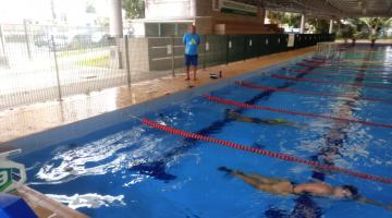 vista geral de piscina com pessoas nadando entre as raias. Homem, do lado de fora, está em pé. #paratodosverem