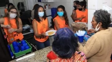 JOvens servem pizza e refrigerante para pessoas atrás de balcão. #paratodosverem