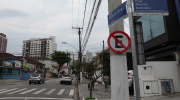 placa de proibido estacionar afixada em poste à direita. Carros passam pela rua. #paratodosverem