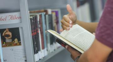 mãos seguram livro aberto. há livros numa estante ao fundo. #paratodosverem 