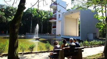 área verdade, com árvore e primeiro plano, banco com quatro pessoas ao lado. Elas estão olhando para um lago com um jato d'água ao centro, que tem, à direita, um mirante de três pavimentos. #paratodosverem