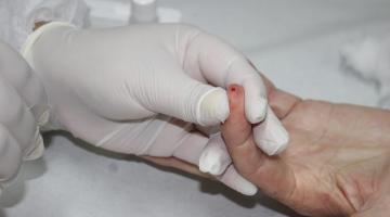 imagem em close de uma mão com luva segurando outra que está com um pequeno furo sangrando, demonstrando um exame de saúde. #paratodosverem