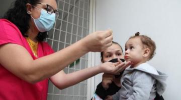 Criança, no colo da mãe, recebe dose de vacina por profissional de saúde. #pratodosverem