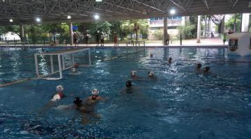 jovens disputam jogo na piscina. Do lado esquerdo e ao fundo se vê a trave sobre a água. #Paratodosverem