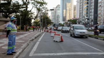 trecho de via interditado com blocos. Carros passam um uma faixa. Agente da CET monitora o trânsito. #paratodosverem