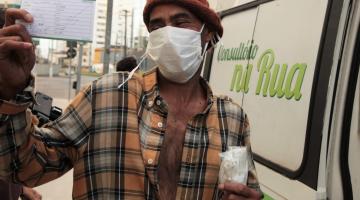 homem exibe cartão de vacinação. Ele está em pé ao lado de uma van onde se lê na porta; Consultório na Rua