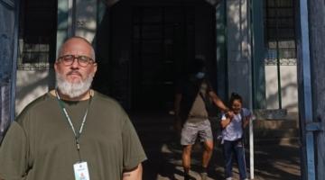 o porteiro Carlos na frente da escola. Há uma criança acompanhada do pai ao fundo saindo da unidade. #paratodosverem