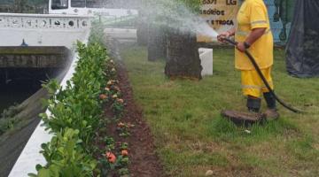 homem uniformizado esguicha água de mangueira em canteiro com flores à margem de um canal. Ele está sobre a grama. #paratodosverem