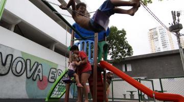 criança está brincando no balancê. Ao fundo, um escorregador com uma mulher segurando uma criança. #paratodosverem