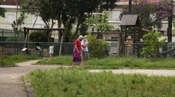 praça com área gramada. Dois idosos caminham. Um playground ao fundo. #paratodosverem