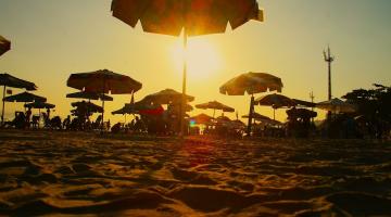 guarda-sois abertos na faixa de areia com o sol ao fundo. #paratodosverem 