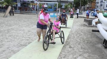 Jovem anda de triciclo, guiada por mulher em pé, em tapete sobre a faixa de areia. #pracegover