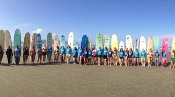 Várias mulheres em pé na areia à frente de seus respectivos long-boards. #Pracegover