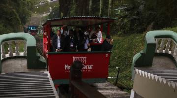 bondinho do monte serrat está em movimento de subida com vários passageiros. Há vegetação do morro ao fundo. #paratodosverem