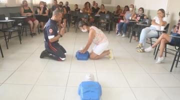 enfermeiro do Samu e uma mulher estão ajoalhados no chão. Entre eles, um boneco serve de simulação para primeiros socorros. Ela aperta o peito do boneco. Outras pessoas sentadas assistem. #paratodosverem