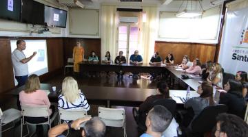 várias pessoas reunidas em torno de uma mesa em forma de U. Duas pessoas estão em pé falando ao grupo. Em uma das paredes está projetada uma imagem. #paratodosverem