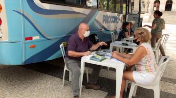 ônibus do procon parado na praça e um homem atendendo uma mulher, com ambos sentados à mesa. #paratodosverem
