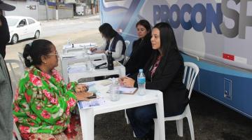 atendentes do procon estão sentadas à frente de van. Uma delas atende uma mulher. #paratodosverem