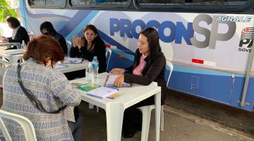 atendentes estão sentadas diante de uma ônibus onde se lê procon na lateral. Uma mulher, de costas para a foto, está sendo atendida. #paratodosverem