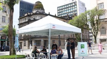 tenda armada em praça com banner do procon ao lado. Há pessoas sentadas. #paratodosverem