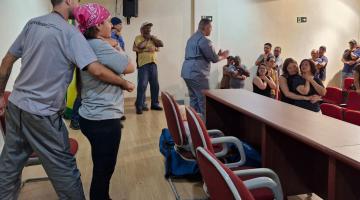 pessoas em pé em auditório simulando desengasgo de pessoa adulta. Um homem está orientando as duplas. #paratodosverem 
