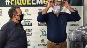 Dois homens estão à frente de pacotes com protetores faciais. Um deles está colocando o protetor na cabeça para demonstrar. Atrás de ambos há um banner onde se lê Fique em Casa. #Paratodosverem