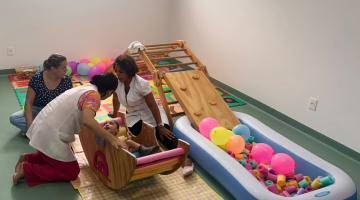 criança com comprometimento de desenvolvimento está dentro de um balanço apoiado no chão e ao lado de peças coloridas. Sob acompanhamento de três mulheres. #paratodosverem 