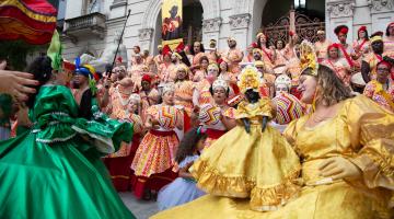 grupo quiloa fantasiado com roupas de desfile de carnaval coloridas dança na escadaria da prefeitura. #paratodosverem 