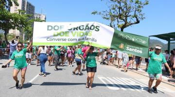 pessoas caminham em avenida segurando faixa. #paratodosverem