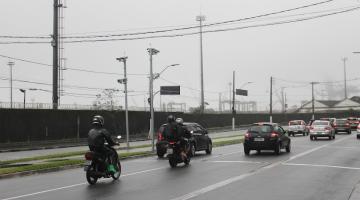 pista de avenida com motos e carros passando. Ao fundo, postes com radares no topo. #paratodosverem