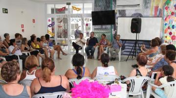 O compositor e comentarista está ao fundo, sentado, e conversa com grupo de idosos em uma grande sala. #paratodosverem