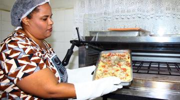 Cristiane está com luvas nas mãos. Ela retira assadeira do forno. #Pracegover