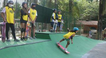 meninas estão na beira da pista aguardnado para ingressar. Uma delas já desliza com o skate na descida da rampa. A área em torno tem bastante vegetação. #paratodosverem