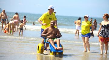 Homem está sentado em cadeira acessível ao mar. Outro homem dirige a cadeira. #Pracegover