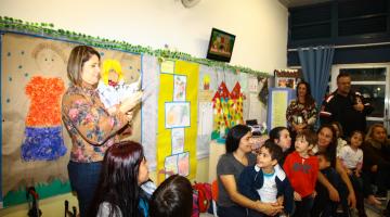 Professora, pais e alunos reunidos em uma sala de educação infantil. A educadora está fazendo uma apresentação aos pais. A parede da sala tem várias colagens de  motivos infantis. #Pracegover