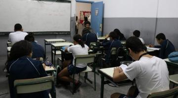 Alunos estão em sala de aula, sentados escrevendo em papéis. Todos estão de costas para a imagem. #Pracegover