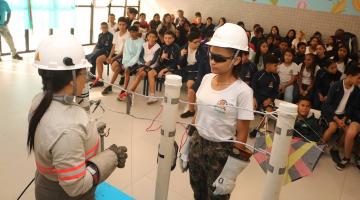 mulher uniformizada e de capacete usa maquete de fiação elétrica para demonstrar riscos com a rede elétrica. Menina à frente, com capacete e óculos, recebe orientação. #paratodosverem 