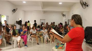 mulher está em pé com livro aberto e lendo para um grupo de outras mulheres que estão sentadas. #paratodosverem