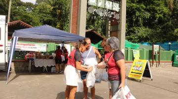 pessoas seguram sacos com doações na porta do orquidário. Ao fundo há uma tenda onde estão concentrados pacotes. #paratodosverem