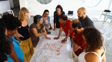 Grupo de pessoas em pé em torno de uma mesa observa mapa da Cidade. #Pracegover