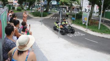 adultos próximos a um muro observam criança dentro de um minibug que passa na pista da praça. #paratodosverem 