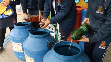 alunos uniformizados despejam óleo de garrafas pet para três galões. #paratodosverem