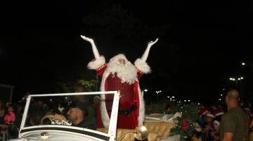papai noel ergue os braços dentro de um carro aberto cumprimentado o público. O carro é dirigido por um motorista. É noite. #paratodosverem 