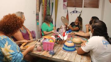 professora ao fundo segura peça artesanal e conversa com alunas, todas sentadas em torno de uma mesa. #paratodosverem 