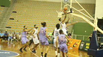 jogo de basquete. Um jogador salta com a bola em direção à cesta, bem próximo à ela. Outros jogadores estão em volta. #paratodosverem