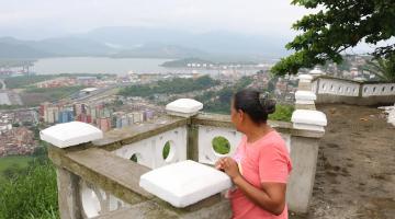 mulher está atrás de mureta no alto de morro olhando para a parte de baixo da cidade, com diversas construções, e o porto ao fundo. #paratodosverem