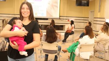 em primeiro plano, mãe segura bebê no colo em pé e sorrindo para a a foto. Ao fundo, uma roda com mulheres conversando. #paratodosverem 