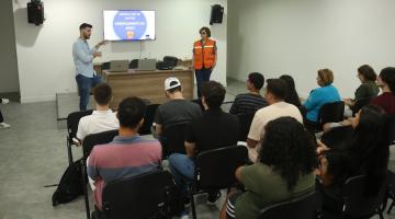 auditório com estudantes sentados e duas pessoas apresentando palestra. #paratodosverem