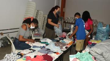 Várias pessoas realizam triagem de roupas que estão em cima da mesa. #paratodosverem